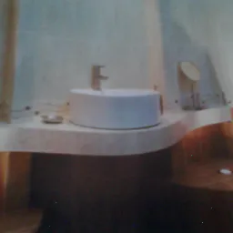 Okrągła, biała umywalka nablatowa z baterią, zamontowana na jasnym, nieregularnym blacie w łazience z beżowymi akcentami i dekoracyjną zasłoną prysznicową.